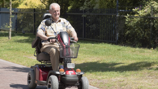 Elderly man riding a red mobility scooter along a sunny park path, smiling and enjoying an outdoor stroll