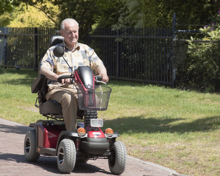Elderly man riding a red mobility scooter along a sunny park path, smiling and enjoying an outdoor stroll