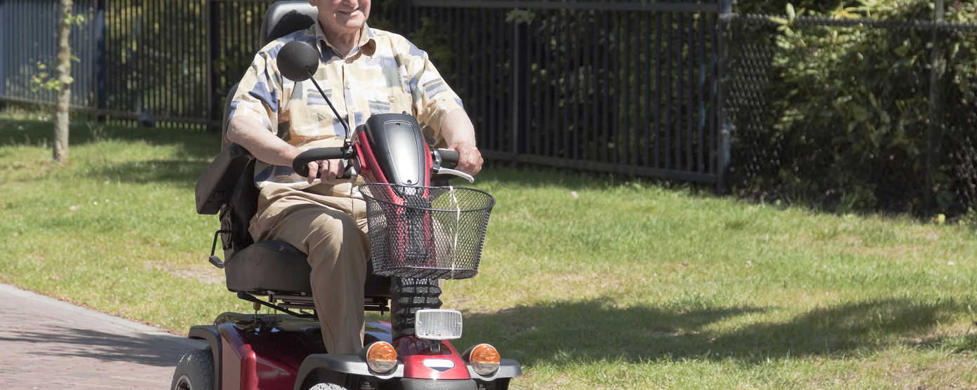 Elderly man riding a red mobility scooter along a sunny park path, smiling and enjoying an outdoor stroll