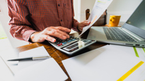 A man in a red plaid shirt is seated at a wooden desk, working on his finances. He is using a large calculator with one hand while holding a printed financial document in the other.