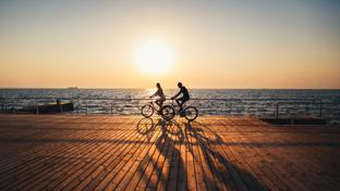 Two cyclists ride along a seaside boardwalk at sunset, casting long shadows over wooden planks