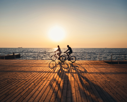 Two cyclists ride along a seaside boardwalk at sunset, casting long shadows over wooden planks