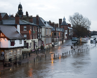 A street next to a river in a historic town is heavily flooded, with water covering the road and sidewalk areas.