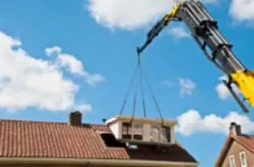A large yellow crane is lifting a prefabricated dormer window structure, onto the roof of a house with red tiles