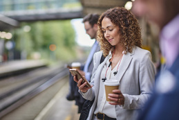 business woman looking at mobile phone at train station with coffee in hand