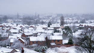 A wide view of a suburban neighborhood blanketed in snow, with rows of houses topped with white rooftops.