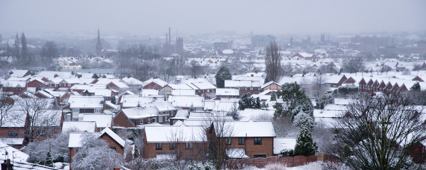 A wide view of a suburban neighborhood blanketed in snow, with rows of houses topped with white rooftops.