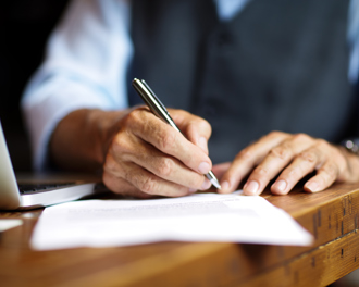 A close-up of hands writing on paper with a pen, a laptop visible in the background.