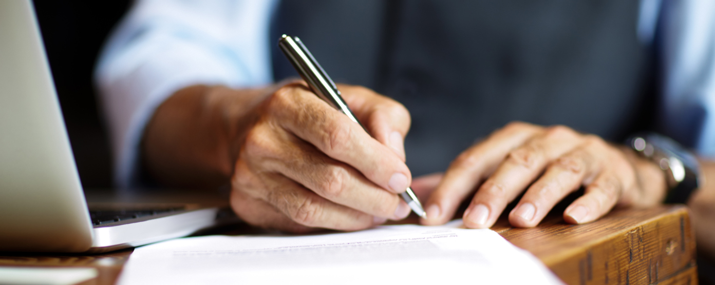 A close-up of hands writing on paper with a pen, a laptop visible in the background.