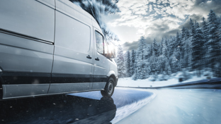 White delivery van navigating a snowy, curving road through a winter forest with blowing snow and low sun