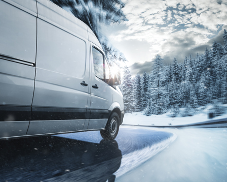 White delivery van navigating a snowy, curving road through a winter forest with blowing snow and low sun