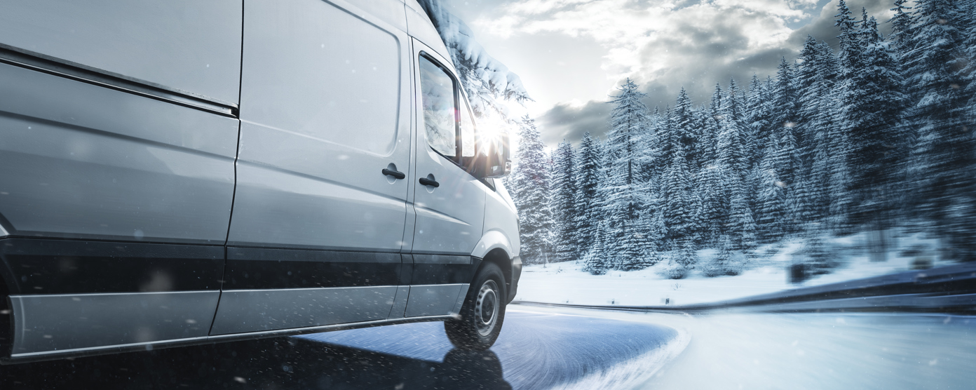 White delivery van navigating a snowy, curving road through a winter forest with blowing snow and low sun