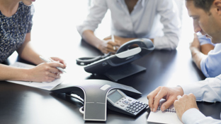 Three business professionals are seated around a conference table, writing notes while participating in a meeting.