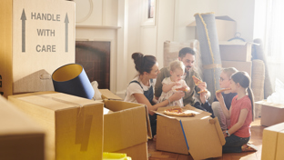 A family unpacks moving boxes in their new home, with a mother holding a toddler, two children excitedly looking at stuffed toys, and a man carrying a rolled-up carpet in the background.