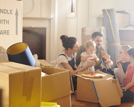 A family unpacks moving boxes in their new home, with a mother holding a toddler, two children excitedly looking at stuffed toys, and a man carrying a rolled-up carpet in the background.