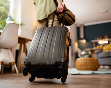 A person is seen from behind pulling a wheeled suitcase and carrying a small travel bag, preparing to leave a modern living space.