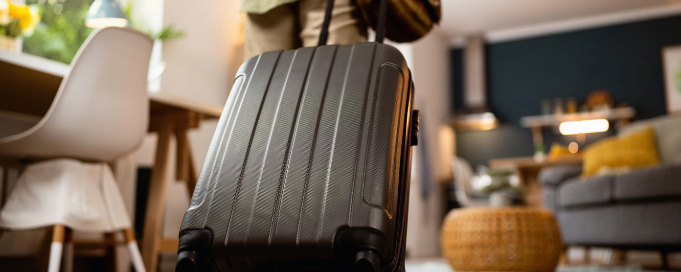 A person is seen from behind pulling a wheeled suitcase and carrying a small travel bag, preparing to leave a modern living space.