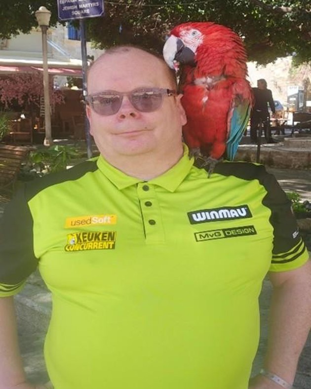 A man stands with his hands on his hips with a green t shirt on and a parrot on his shoulder