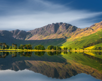 Mountain with calm waters reflecting the surrounding mountains and a line of trees along the shore under a clear sky