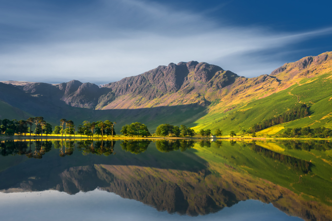 Mountain with calm waters reflecting the surrounding mountains and a line of trees along the shore under a clear sky