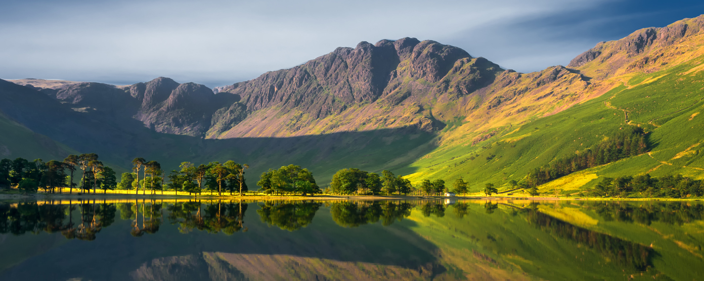 Mountain with calm waters reflecting the surrounding mountains and a line of trees along the shore under a clear sky