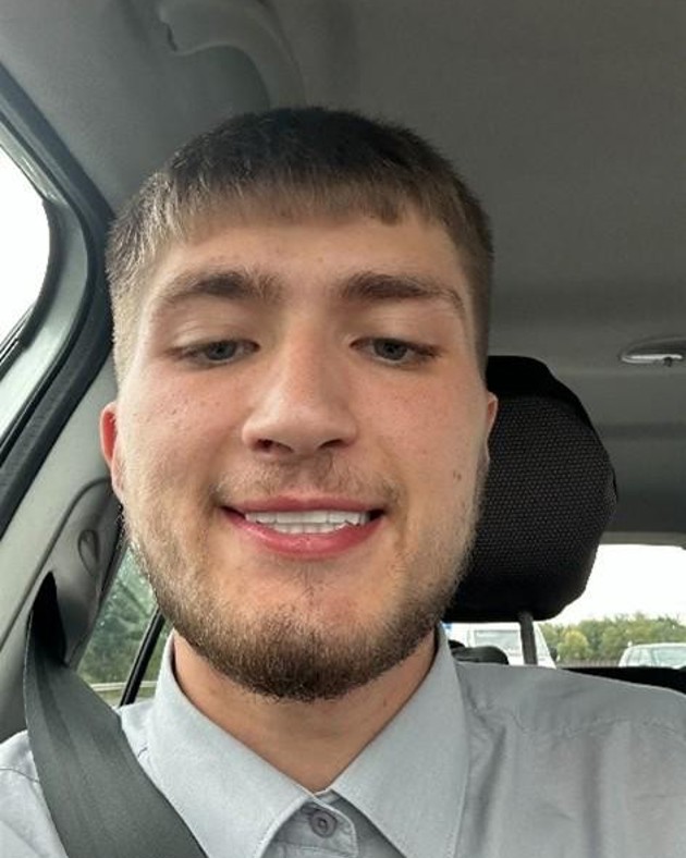 A young man wearing a grey collared shirt sits smiling in his car 