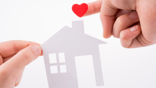 Close-up of two hands holding a white paper cut-out of a house, with a small red heart on one fingertip.
