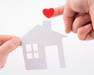 Close-up of two hands holding a white paper cut-out of a house, with a small red heart on one fingertip.