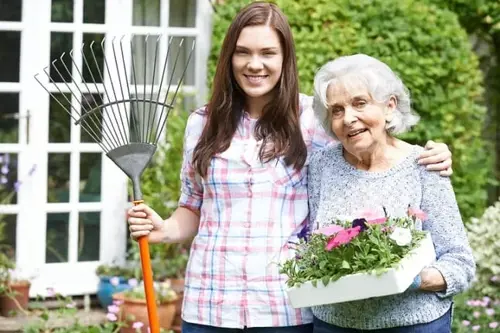 Smiling young woman and her grandmother gardening together in a backyard, holding a rake and a tray of colorful flowers in front of a house