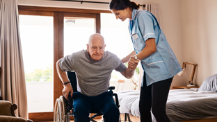 A female caregiver helping an elderly man rise from a wheelchair in a bright, comfortable room, demonstrating support and compassion in a caregiving environment