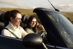 Two women smiling and enjoying a drive in a convertible car under a bright, sunny sky.