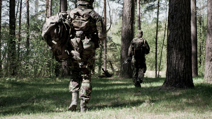 Two soldiers in camouflage gear walking through a sunlit forest with tall trees.
