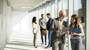 Senior executive and colleague reviewing a tablet in a bright office corridor, with coworkers talking in the background