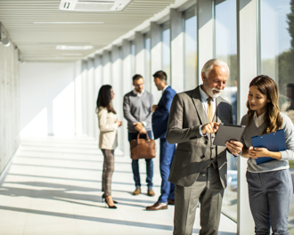Senior executive and colleague reviewing a tablet in a bright office corridor, with coworkers talking in the background