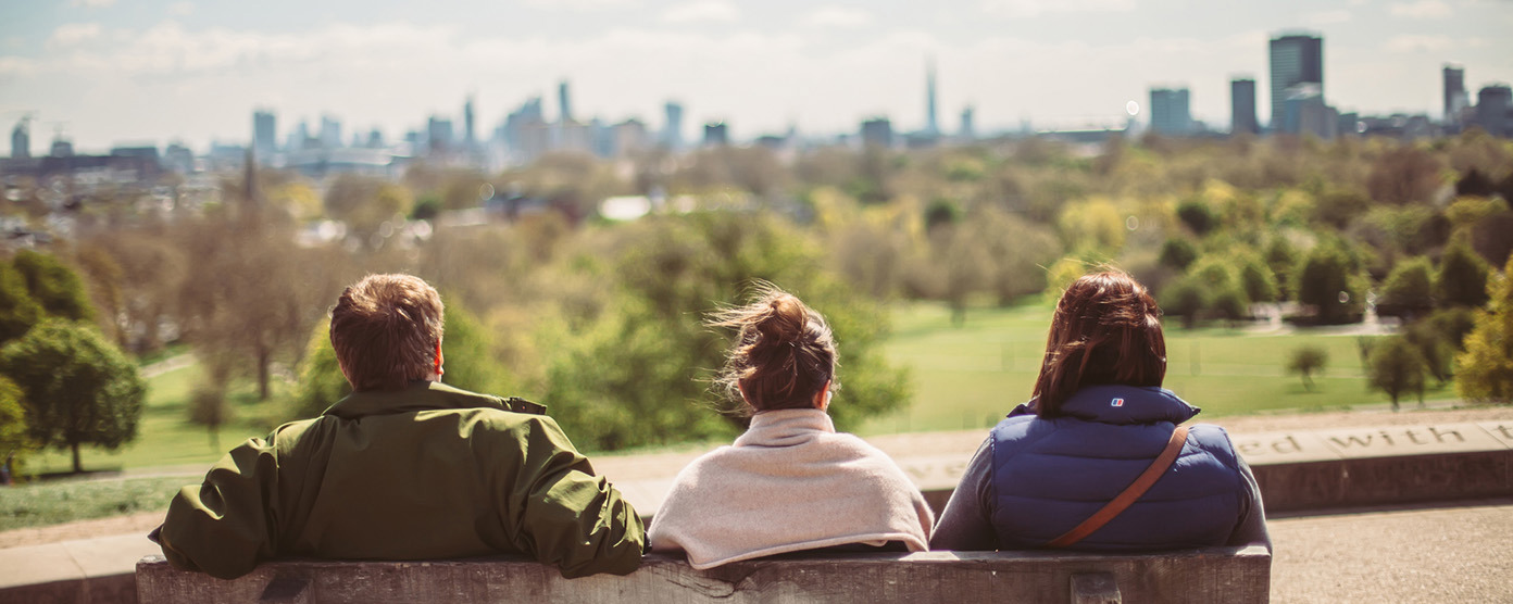 3 on bench, in a park with trees and busy towers along the skyline (Primrose Hill in North London, overlooking the city)