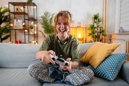 A young woman smiles at the camera as she plays with a game console on her sofa