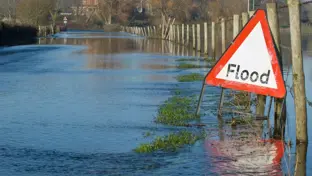 Red triangular flood warning sign beside a submerged country road with water covering the lane and fields