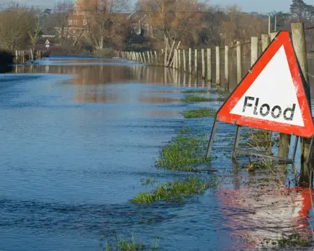 Red triangular flood warning sign beside a submerged country road with water covering the lane and fields
