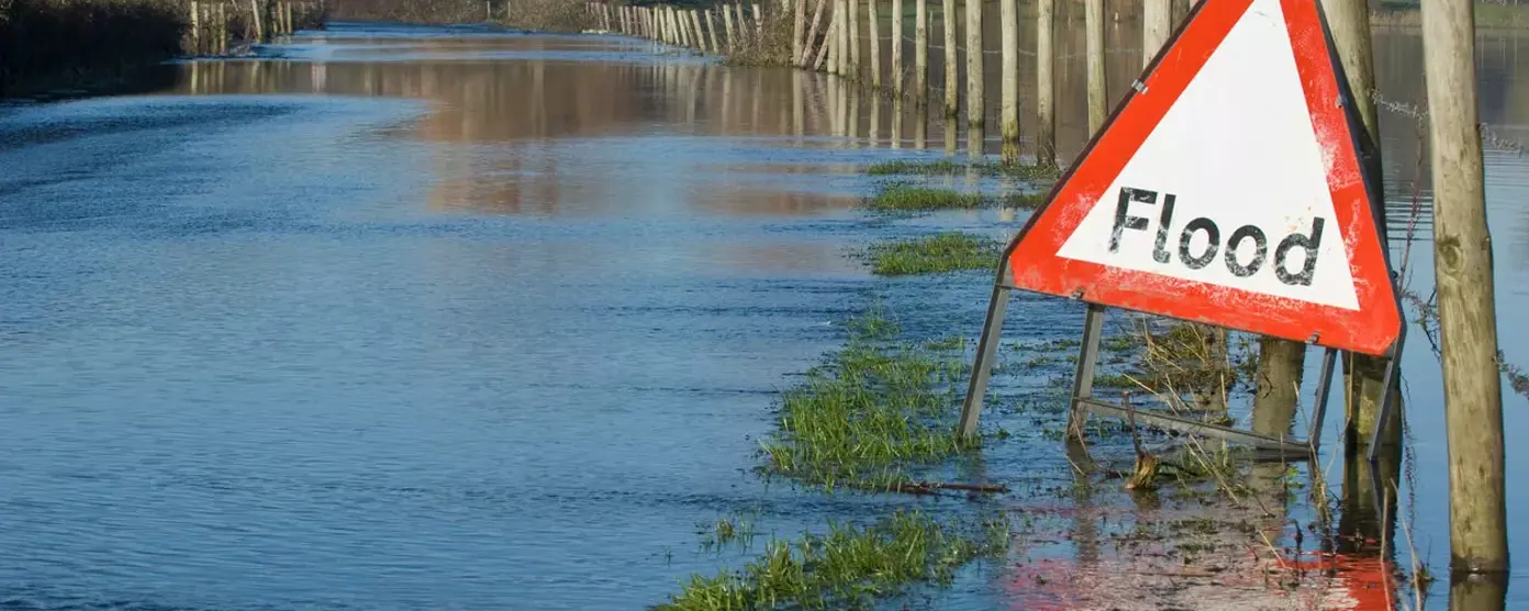 Red triangular flood warning sign beside a submerged country road with water covering the lane and fields
