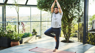 Man practicing yoga in tree pose on a mat in a bright, plant-filled balcony space