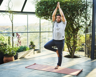 Man practicing yoga in tree pose on a mat in a bright, plant-filled balcony space