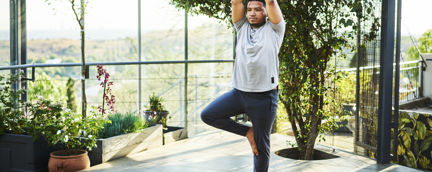 Man practicing yoga in tree pose on a mat in a bright, plant-filled balcony space