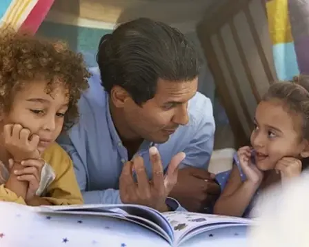 A father and his two young children, a boy and a girl, are lying on the floor inside a blanket fort, looking at an open book together.