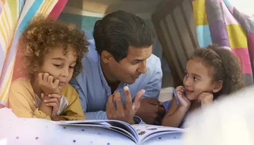 A father and his two young children, a boy and a girl, are lying on the floor inside a blanket fort, looking at an open book together.