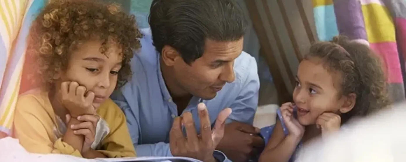 A father and his two young children, a boy and a girl, are lying on the floor inside a blanket fort, looking at an open book together.