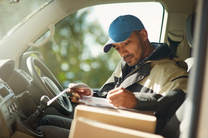 Delivery driver in a van filling out paperwork on a clipboard with parcels on the seat