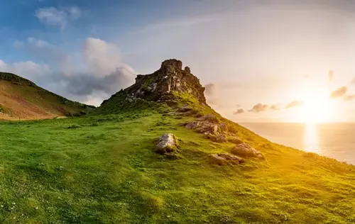 A dramatic rocky hilltop covered in green grass, sloping down toward the ocean, illuminated by a vibrant sunset casting golden light across the landscape with scattered clouds in the sky.