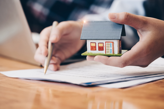 Person holding a small model house while reviewing and signing real estate or property documents at a desk.