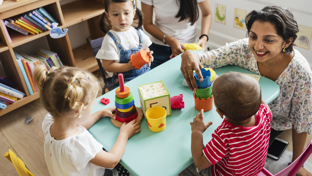 Nursery teacher engaging toddlers with stacking toys at a small activity table in a colorful classroom