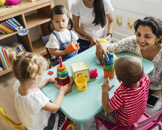 Nursery teacher engaging toddlers with stacking toys at a small activity table in a colorful classroom